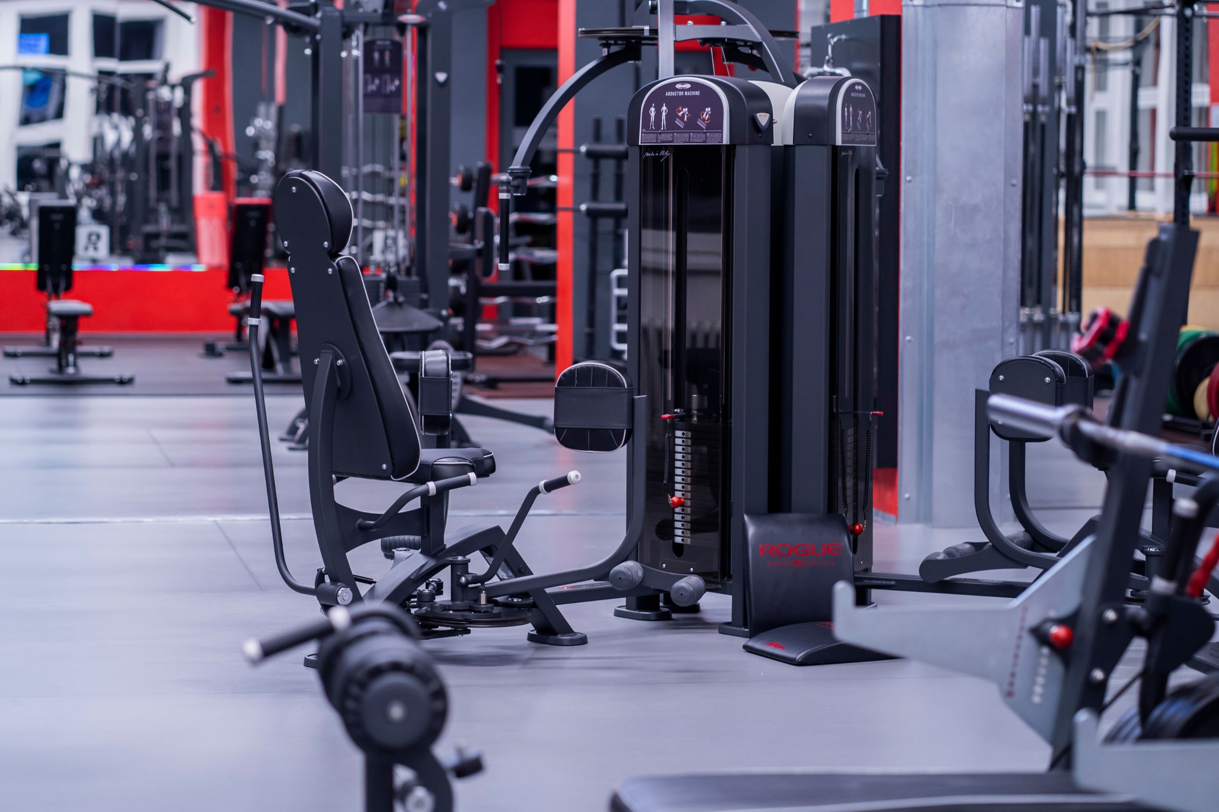 Moderní, prázdná posilovna s šedou podlahou a červenými akcenty na sloupcích. Stropní konstrukce nese žluté popruhy, podél stěn je uspořádáno různé fitness vybavení jako činky a medicinbaly.